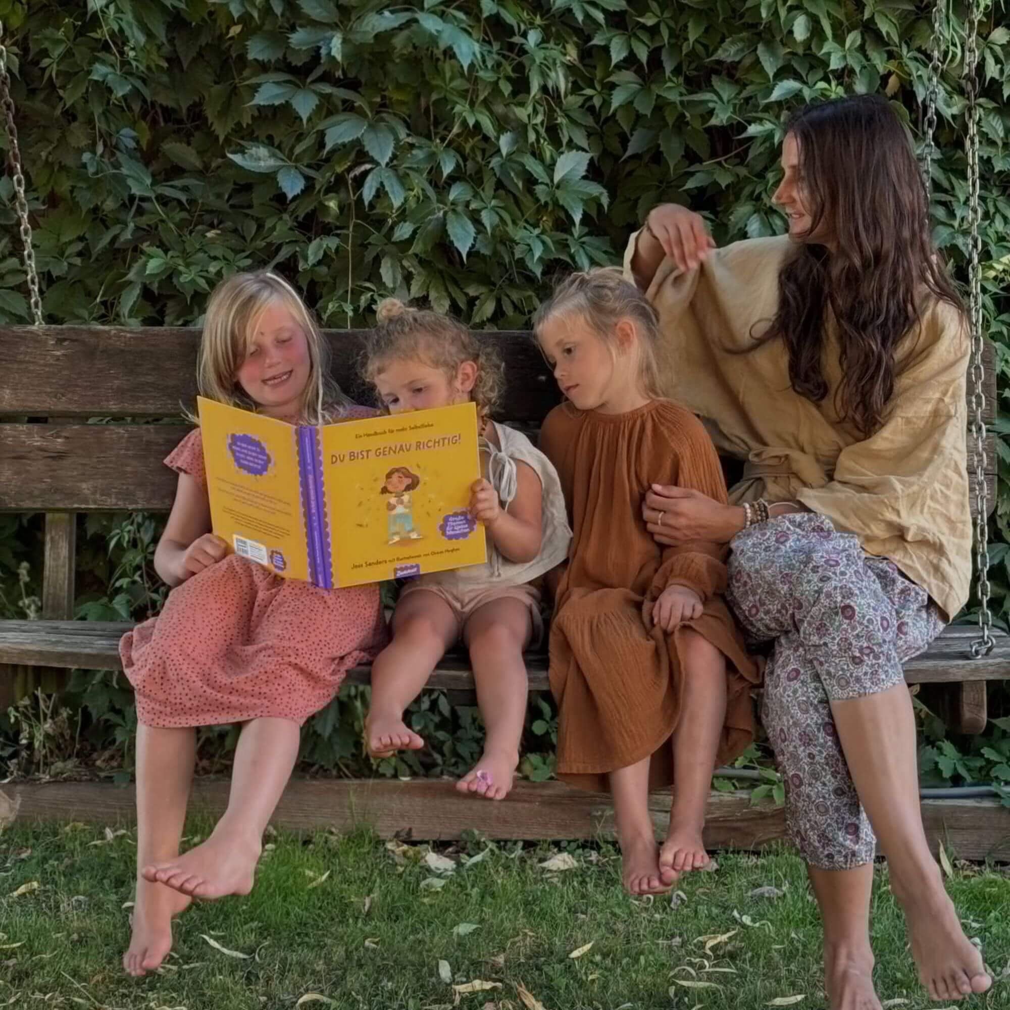 Eine Frau und drei barfüßige Mädchen sitzen auf einer Holzschaukel. Ein Mädchen liest laut aus dem gelben Buch "Du bist genau richtig" von Jessica Sanders vor. Grüne, belaubte Reben im Hintergrund schaffen eine gemütliche Szene im Freien.