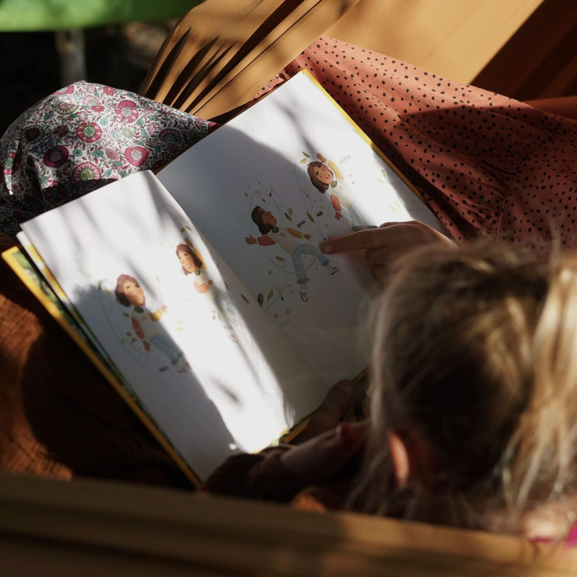 Ein blondes Kind liest "Du bist genau richtig" von Jessica Sanders und zeigt dabei auf eine Illustration. Das Sonnenlicht lässt das Buch und den Stoff schimmern und fängt einen warmen Moment der Selbstliebe und Entdeckung ein.