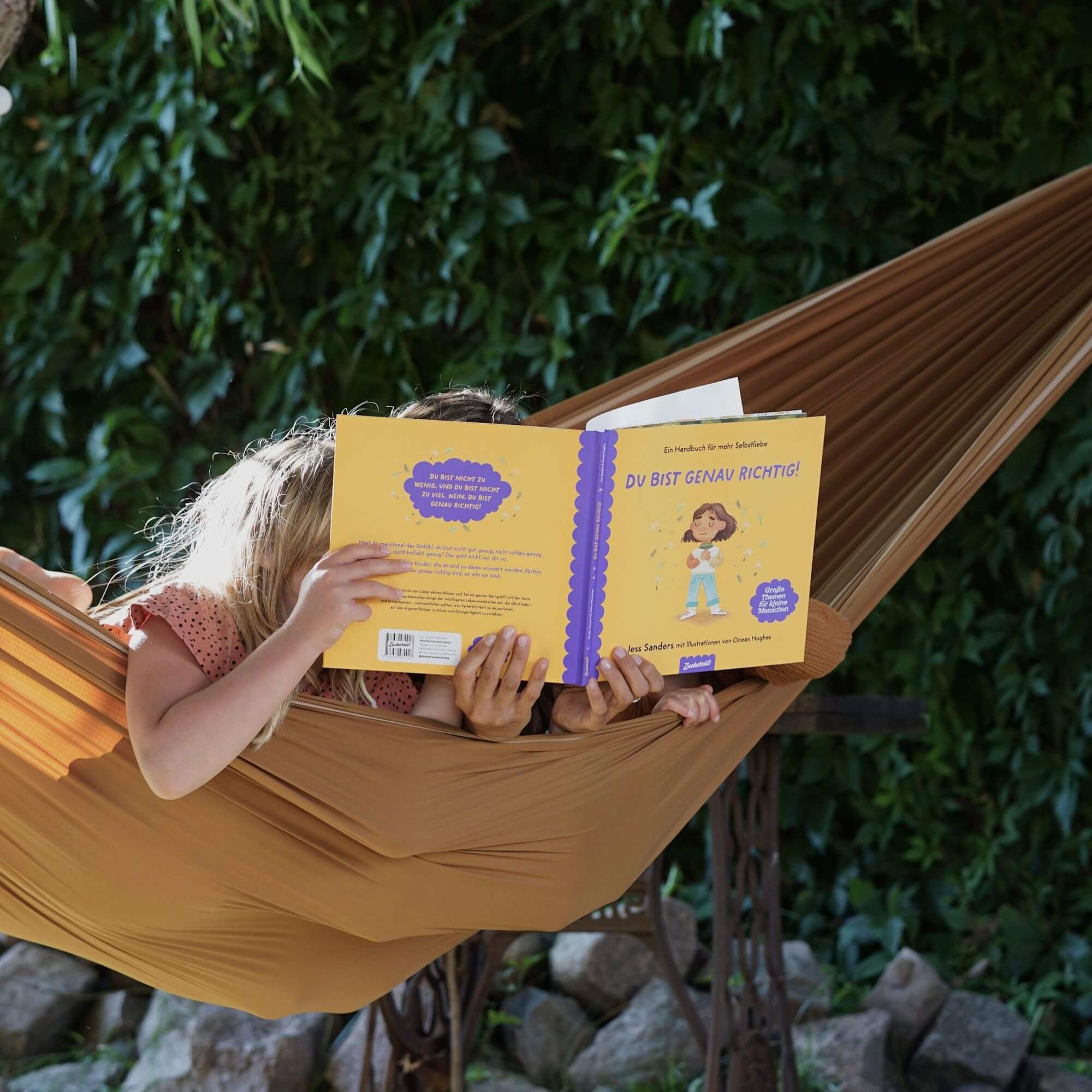 Zwei Kinder entspannen sich im Freien in einer Hängematte und lesen "Du bist genau richtig" von Jessica Sanders. Üppige grüne Pflanzen bilden den Hintergrund, während sie dieses inspirierende Kinderbuch über Selbstliebe genießen.