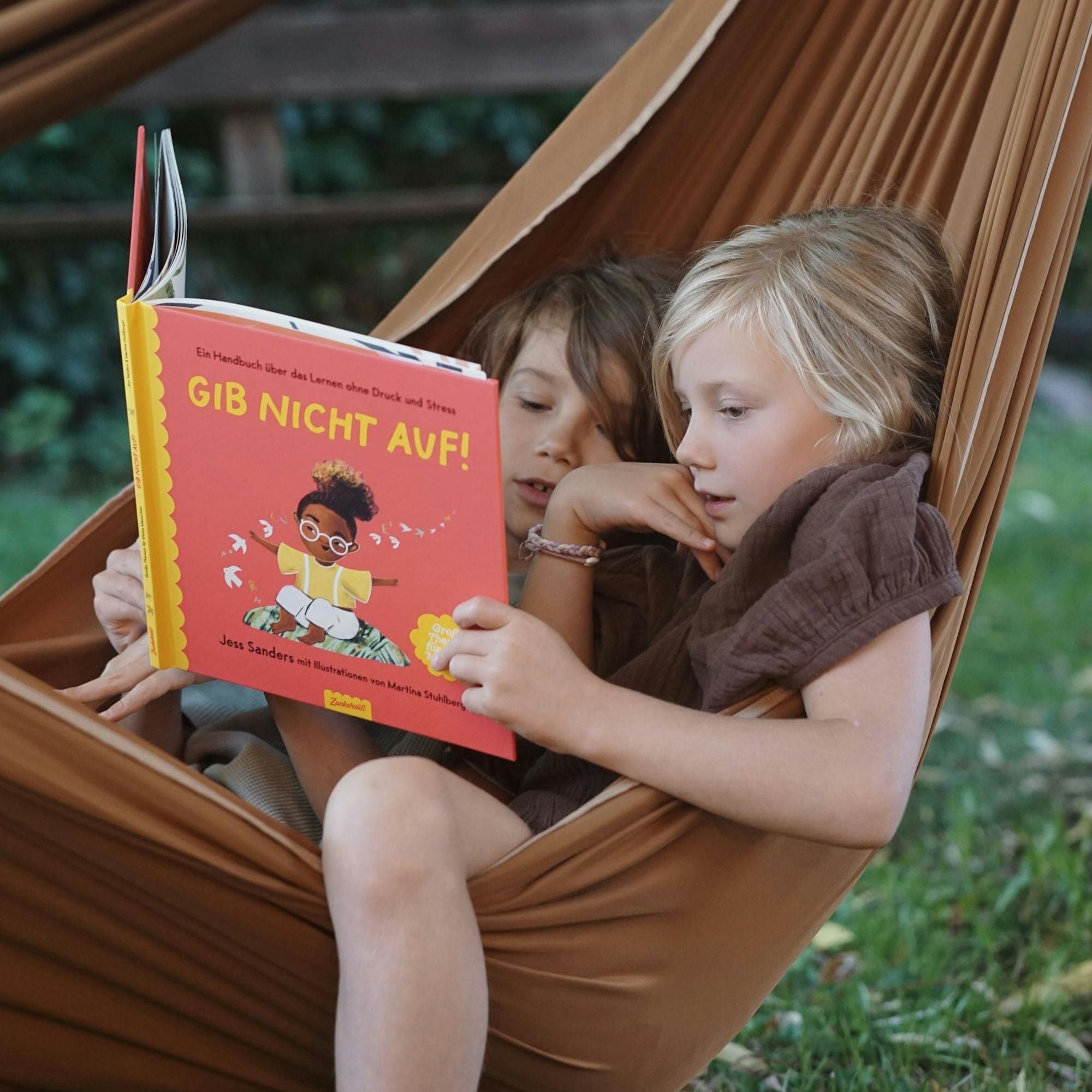 Zwei Kinder lesen das Buch "Gib nicht auf!" von Jess Sanders des Zuckersüß Verlags im Freien. Sie liegen dabei in einer braunen Hängematte. Im Hintergrund ist grünes Gras zu sehen. In dem Buch geht es um Durchhaltevermögen und Selbstvertrauen.