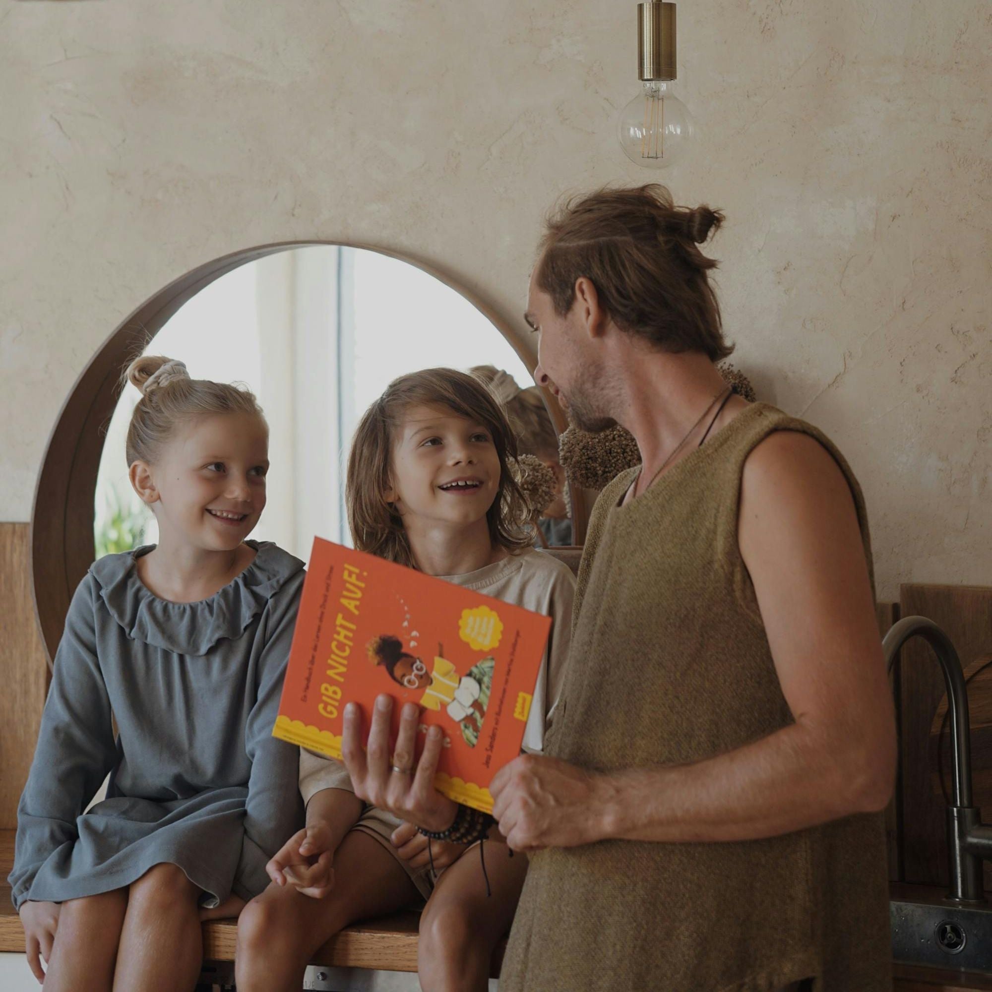 Ein Erwachsener hält das Kinderbuch „Gib nicht auf!“ von Jess Sanders aus dem „School Starter Pack“ des Zuckersüß Verlags in der Hand und unterhält sich lachend mit zwei Kindern, die auf einem Tresen sitzen. Die Szene spielt in einem gemütlich eingerichteten, warm beleuchteten Raum mit rundem Spiegel im Hintergrund. Alle wirken fröhlich und verbunden.