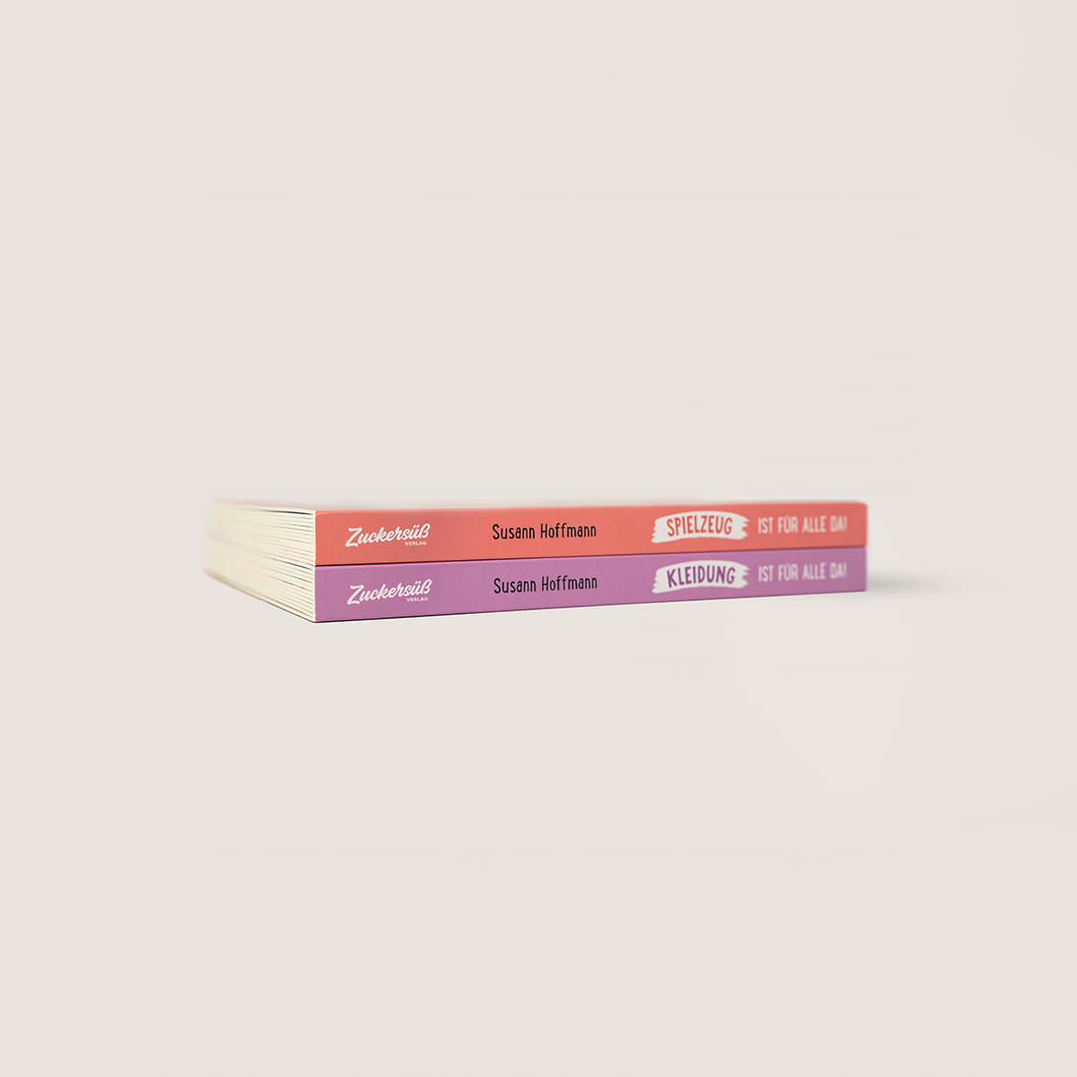 Die Bücher "Spielzeug ist für alle da" und "Kleidung ist für alle da" von Zuckersüß Verlag liegen gestapelt auf heller Fläche, das obere mit rotem, das untere mit pinkem Rücken. Der Hintergrund ist schlicht off-white.