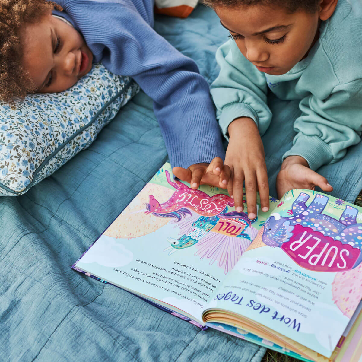 Zwei Kinder liegen auf einem Bett mit blauer Bettwäsche und einem geblümten Kissen und lesen "Entspannt wie ein Panda" von Kira Willey. Ein Kind zeigt auf die Seiten, während beide in die bunten Illustrationen und den Text des Buches vertieft sind.