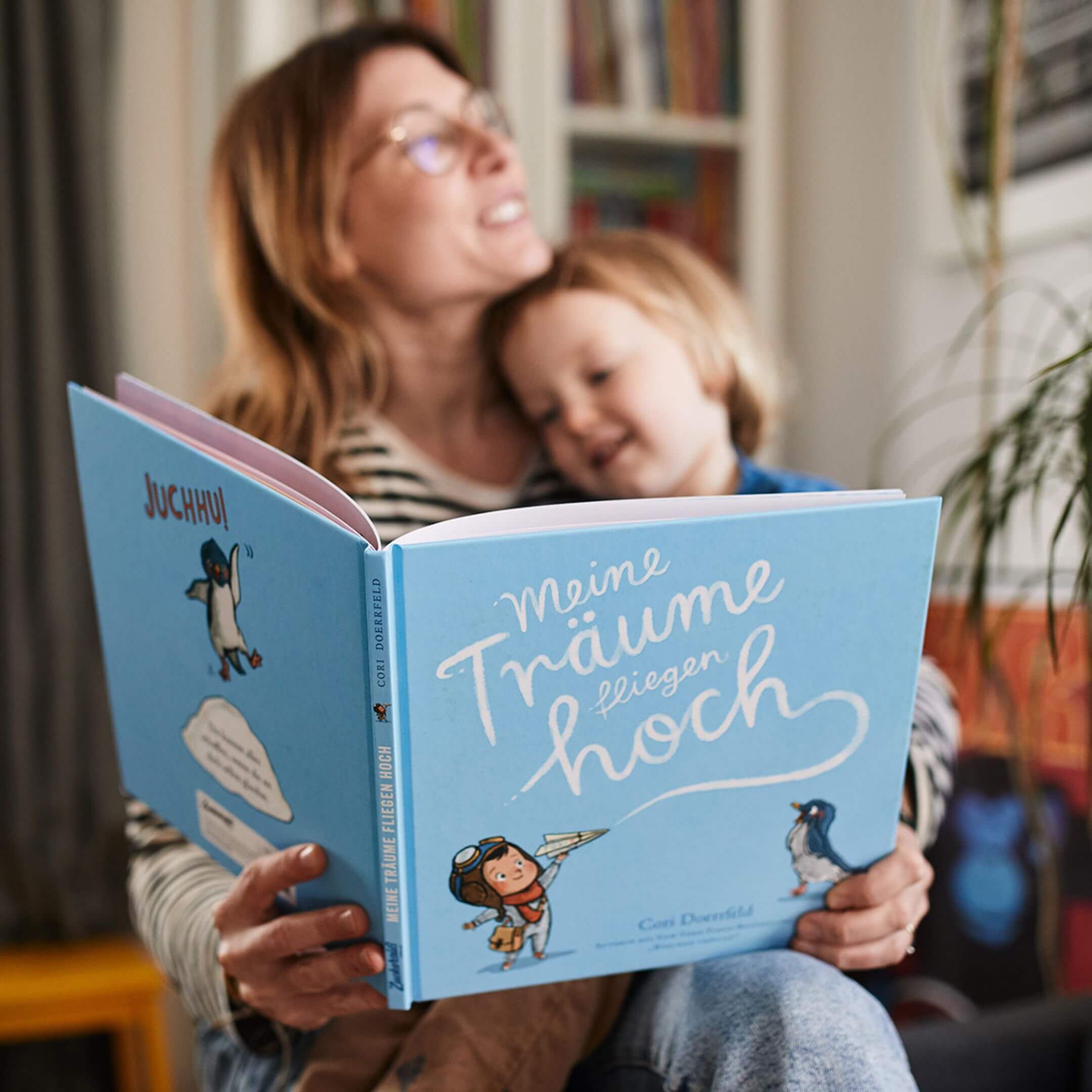Ein lächelnder Erwachsener und ein Kind lesen gemeinsam auf einer Couch ein blaues deutsches Kinderbuch mit dem Titel Meine Träume fliegen hoch, beide wirken glücklich und gemütlich in einem warm beleuchteten Raum.