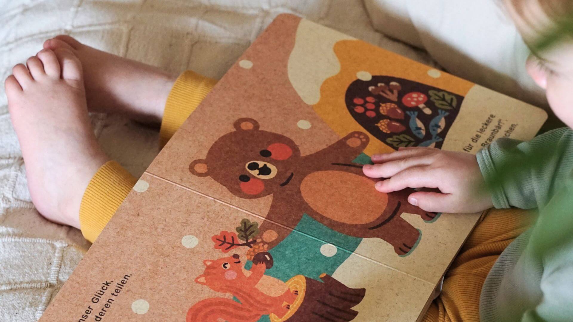 Ein Kind in einer gelben Hose sitzt mit gekreuzten Beinen da und berührt sanft ein Pappbuch mit bunten Illustrationen von einem Bären und einem Eichhörnchen.
