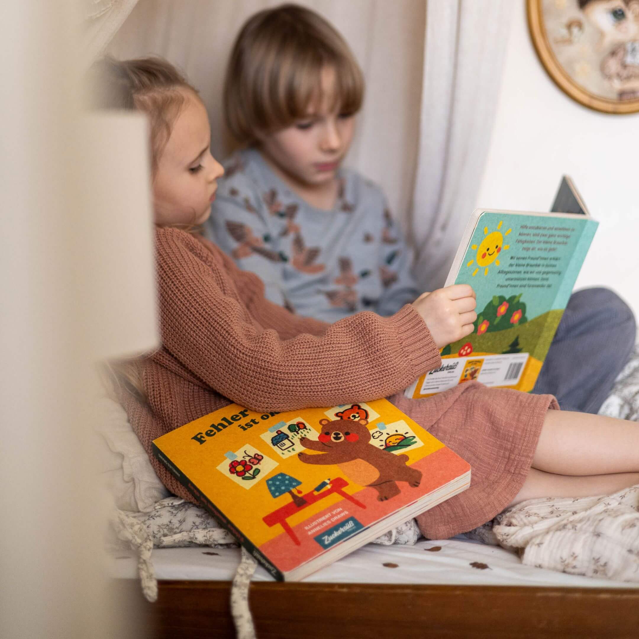 Zwei kleine Kinder kuscheln auf einem gemütlichen Bett und lesen bunte Kinderbücher. Das eine hält "Hilfe brauchen ist okay" in der Hand, das andere "Fehler machen ist okay" von Annelies Draws. 