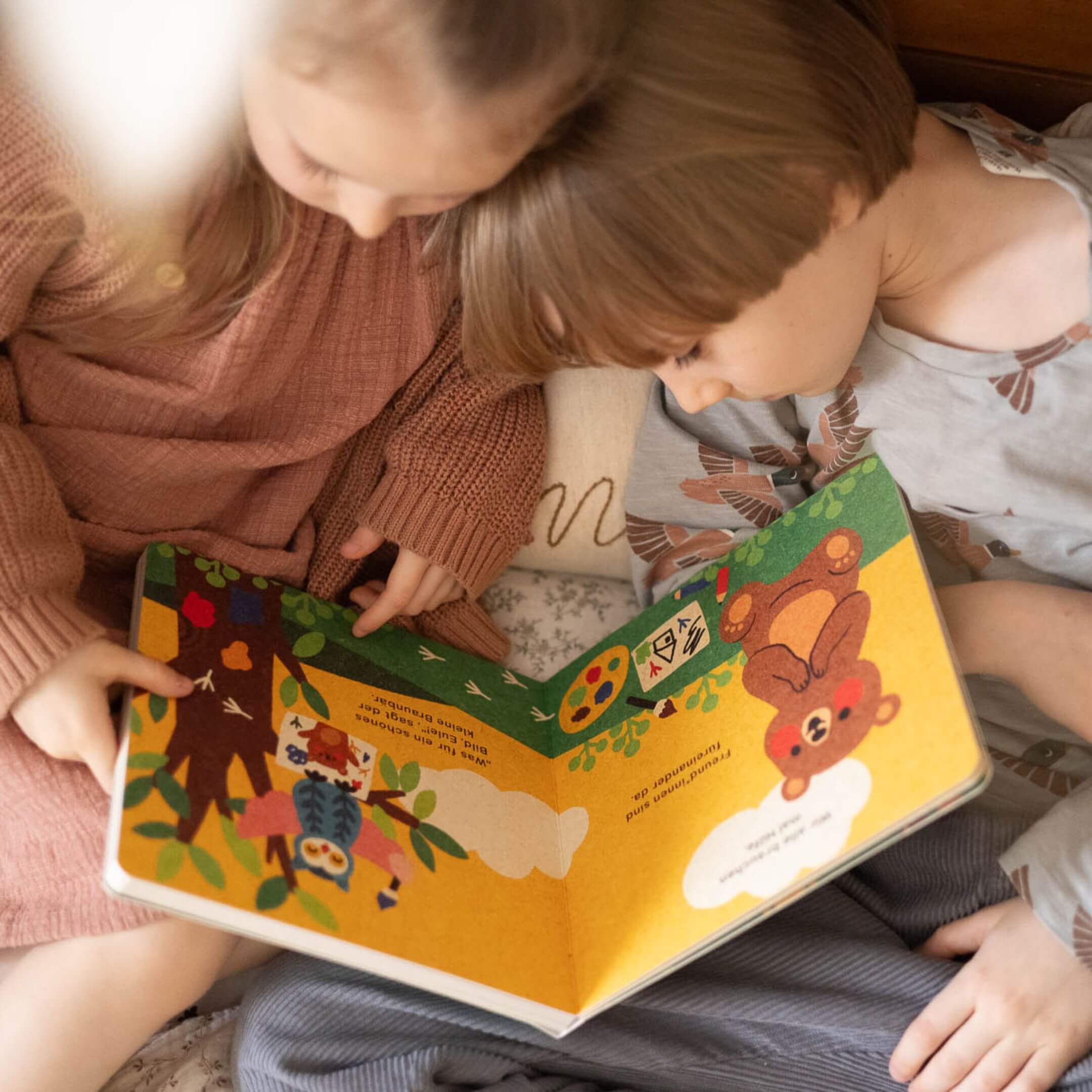 Zwei Kinder sitzen eng nebeneinander auf dem Bett und betrachten gemeinsam eine aufgeschlagene Seite des Bilderbuchs „Hilfe brauchen ist ok“ von Annelies Draws. Die Szene zeigt einen Bären, eine Eule und bunte Naturmotive. 
