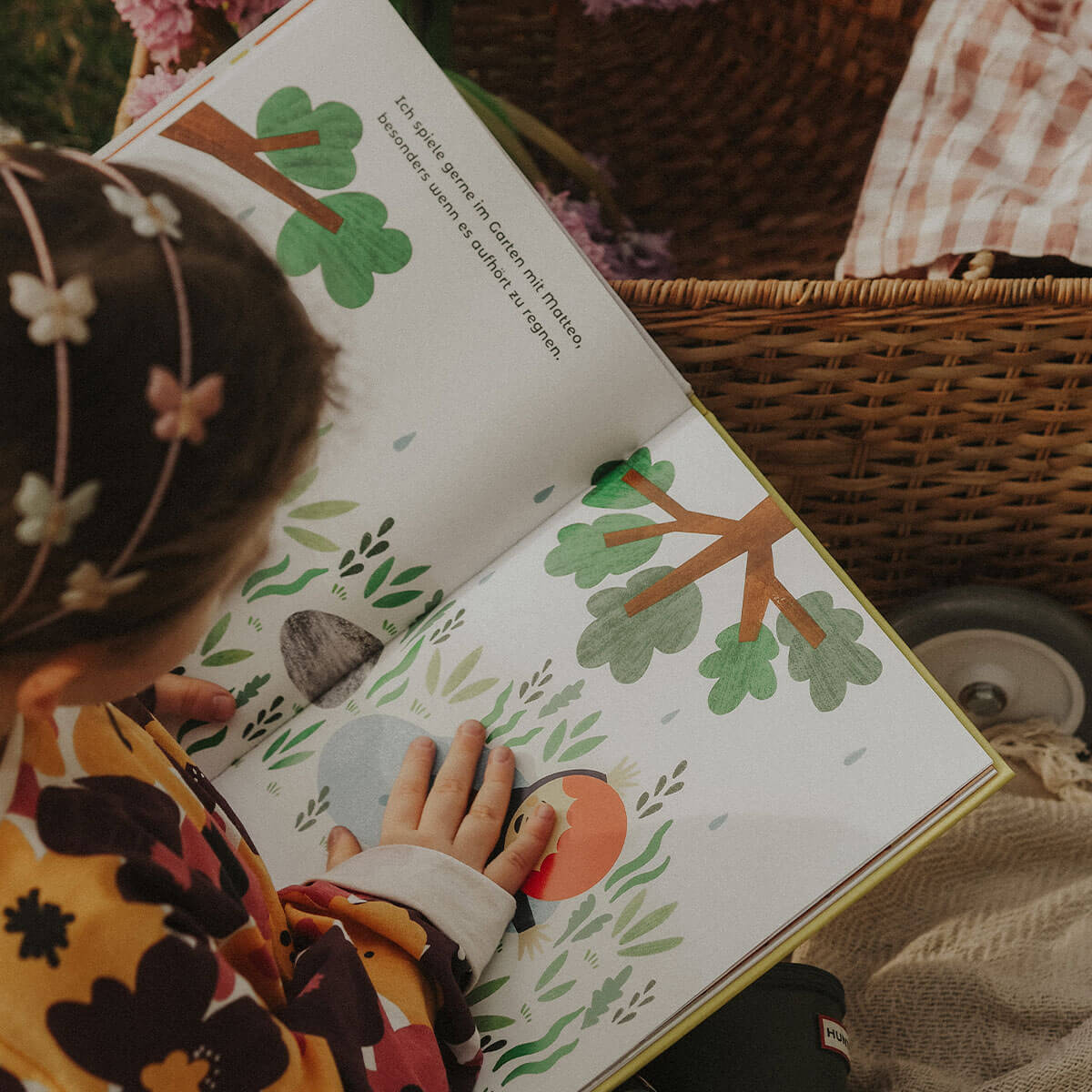 Ein kleines Kind mit einem Blumenstirnband liest im Freien neben einem Weidenkorb 