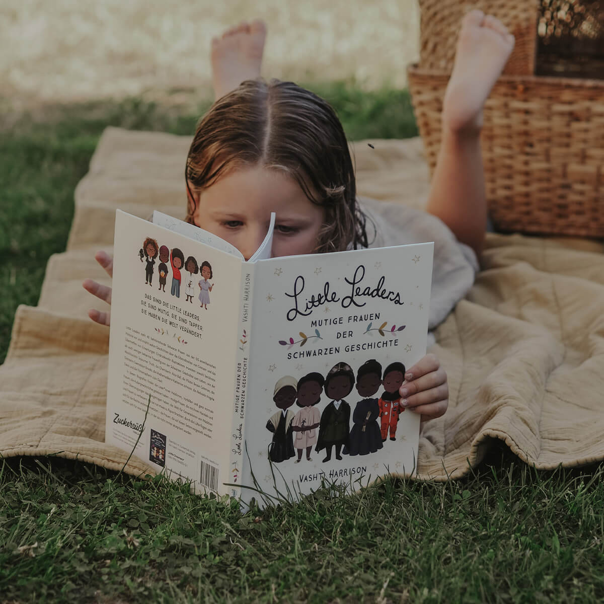Ein kleines Kind liegt draußen auf einer beigen Decke und liest „Little Leaders“ von Vashti Harrison. Im Hintergrund steht ein Weidenkorb.